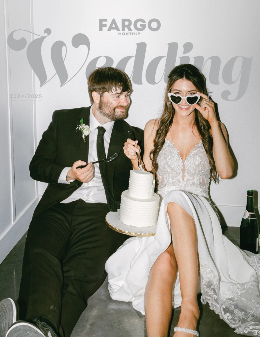 Bride and groom sitting on floor with wedding cake, Fargo Monthly magazine cover 2024/2025. Bride in white dress with heart-shaped sunglasses, groom in black suit, both holding forks over a two-tier white wedding cake.