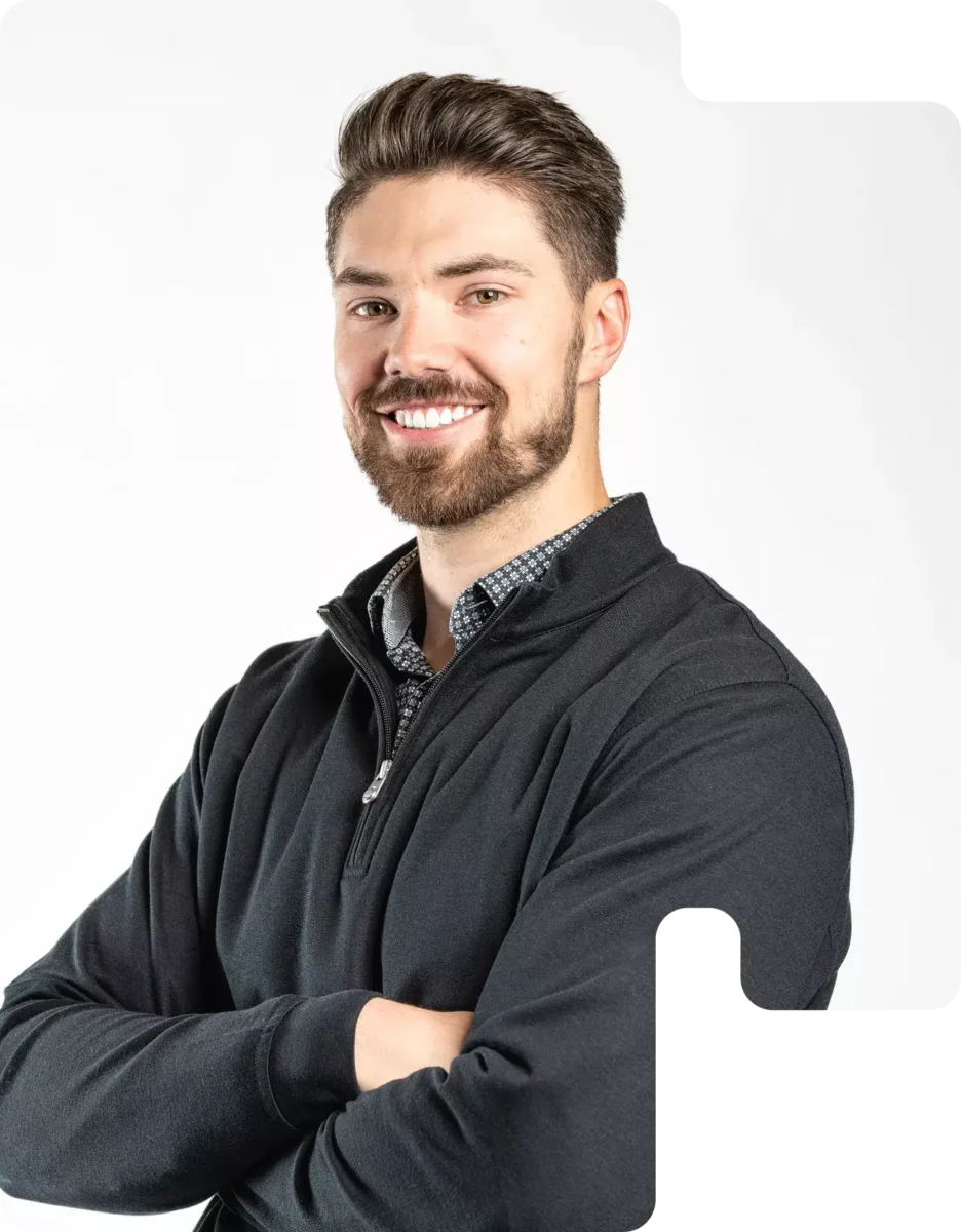 Smiling Man With Folded Arms Wearing A Black Quarter-Zip Pullover And Patterned Shirt On A White Background.